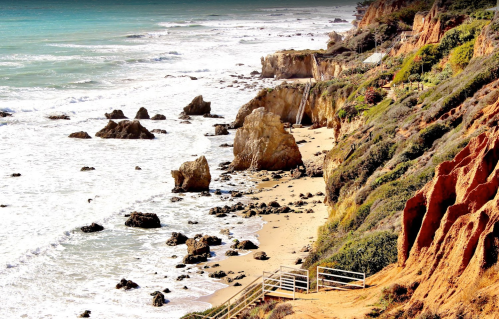 A scenic coastal view with rocky cliffs, sandy beach, and waves crashing against the shore under a clear sky.