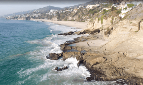 Aerial view of a rocky coastline with waves crashing, sandy beach, and cliffs in the background.