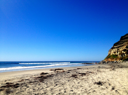 A sunny beach scene with clear blue skies, gentle waves, and a rocky cliff in the background.