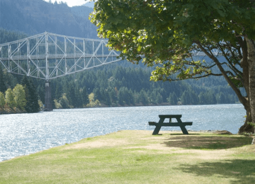 A picnic table by a river, with a bridge in the background and trees lining the shore.