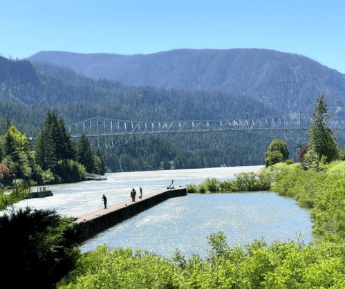 A scenic view of a river with a dock, surrounded by trees and mountains, under a clear blue sky.