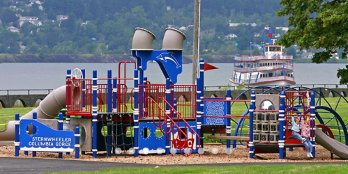 Colorful playground equipment with slides and climbing structures, overlooking a river and a boat in the background.