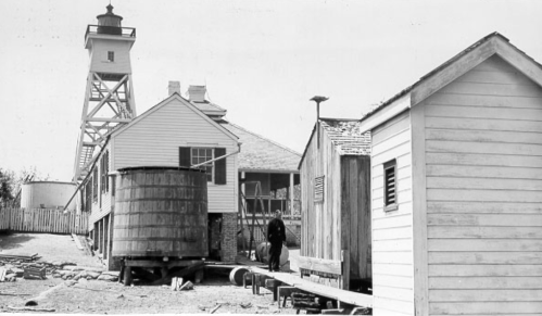 Historic black-and-white photo of a lighthouse, wooden buildings, and a person walking along a path.