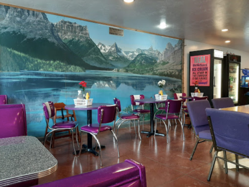 A cozy diner with purple chairs and tables, featuring a scenic mountain mural and ice cream signage on the wall.