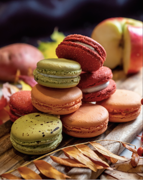 A colorful assortment of macarons stacked on a wooden board, surrounded by autumn leaves and fruits.