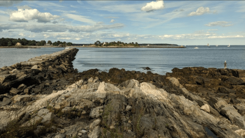 A rocky shoreline extends into calm waters under a cloudy sky, with a person fishing in the distance.