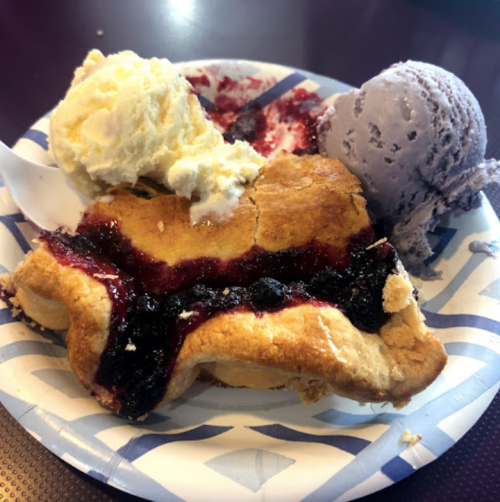 A slice of berry pie topped with vanilla and purple ice cream on a paper plate.