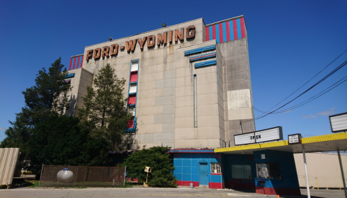 Abandoned Ford-Wyoming Drive-In theater with colorful facade and overgrown landscaping under a clear blue sky.