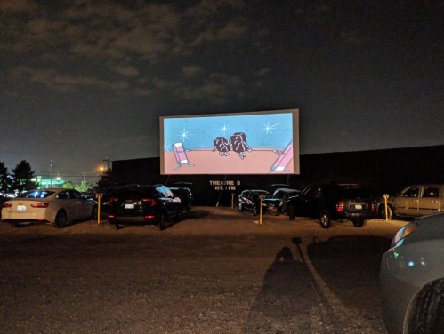 A drive-in theater screen shows an animated scene at night, with parked cars in the foreground.