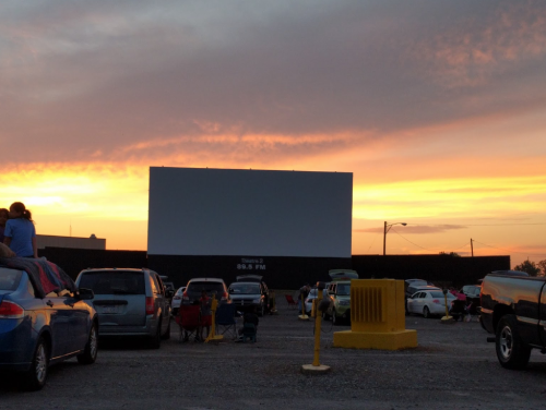 A drive-in theater at sunset, with cars parked and a large blank screen ready for a movie.