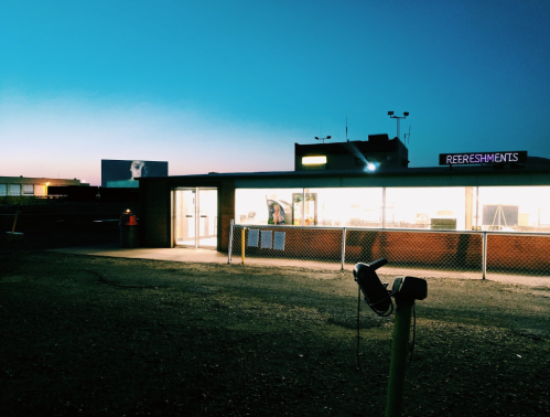 A brightly lit refreshment stand at dusk, with a large billboard in the background and a clear blue sky.