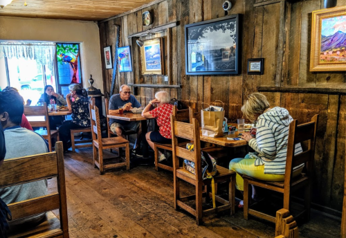 A cozy restaurant interior with wooden walls, featuring diners at tables and colorful artwork on display.