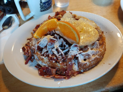 A plate of waffles topped with bacon, powdered sugar, orange slices, and a dollop of cream.
