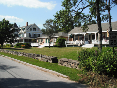A quiet street lined with charming houses and a stone wall, featuring green lawns and a speed limit sign.