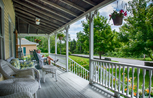 A person relaxes on a porch with rocking chairs, surrounded by greenery and hanging flower pots.