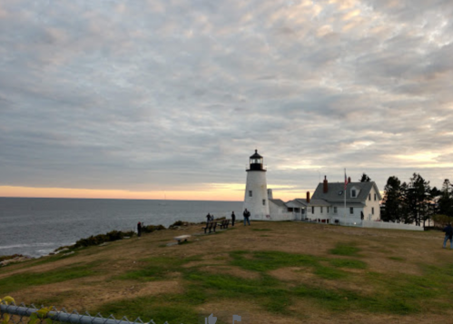 A lighthouse stands on a grassy hill by the ocean, with people nearby and a cloudy sunset sky in the background.