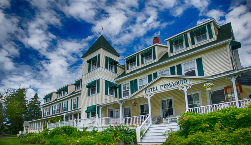 Historic Hotel Pemaquid with a tower, green shutters, and a wraparound porch, set against a blue sky with clouds.
