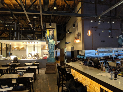 Interior of a modern restaurant featuring a bar, wooden tables, and a mural of a statue on the wall.