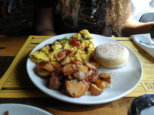 A plate with an omelette topped with tomatoes and herbs, roasted potatoes, and an English muffin.