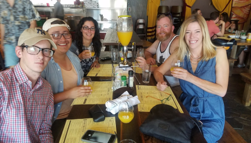 A group of five friends enjoying drinks at a restaurant, smiling and seated around a table with menus and glasses.