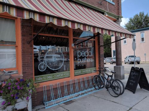 Exterior of a restaurant called "Pedal Pushers," featuring a striped awning, bicycles, and a signboard with menu items.
