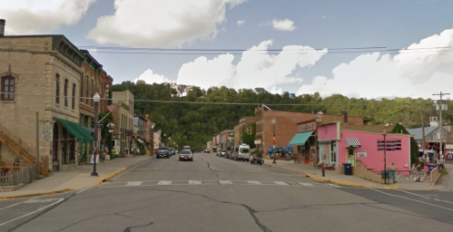 A quaint small town street with shops, cars, and green hills under a partly cloudy sky.