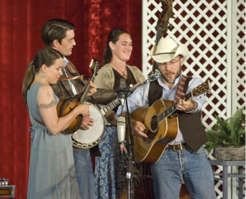 A bluegrass band performs on stage, featuring a guitarist, banjo player, and two women with instruments.