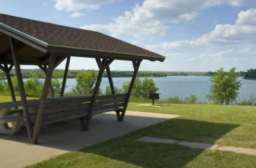 A wooden shelter overlooks a calm lake, surrounded by green grass and trees under a partly cloudy sky.