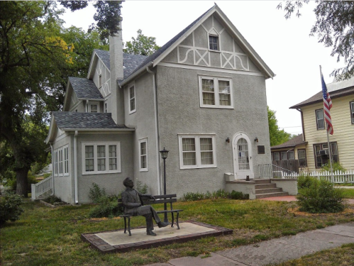 A gray house with white trim and a statue of a seated man on a bench in front, surrounded by greenery. An American flag is visible.