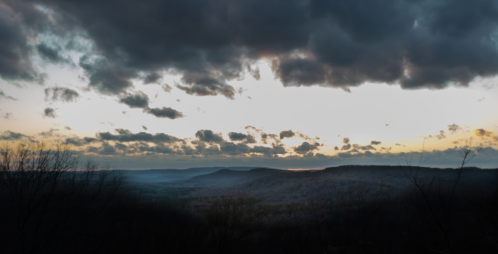 A panoramic view of rolling hills under a cloudy sky at sunset, with mist rising in the valleys.