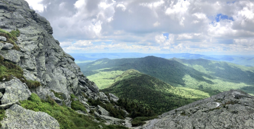 A panoramic view of lush green mountains under a partly cloudy sky, with rocky cliffs in the foreground.