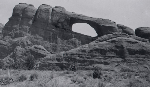 A large rock formation with a natural arch, surrounded by sparse vegetation and rugged terrain.