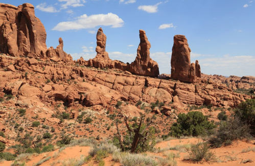 Rock formations rise against a blue sky in a desert landscape, with sparse vegetation and rugged terrain.