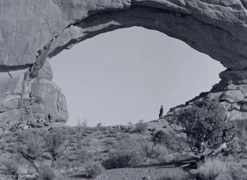 A person stands beneath a large rock arch in a desert landscape, surrounded by sparse vegetation and rocky terrain.