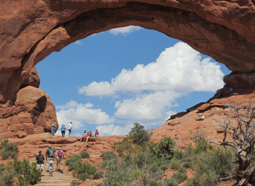A natural rock arch frames a group of hikers exploring a desert landscape under a blue sky with fluffy clouds.