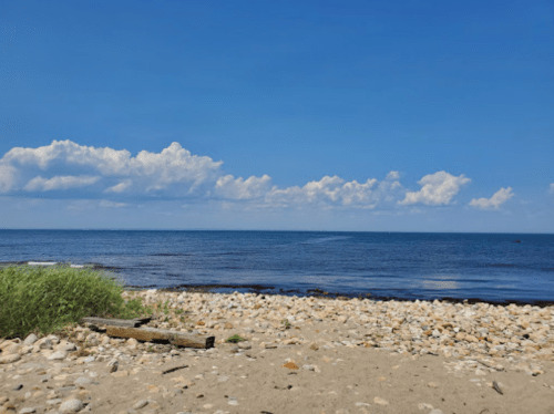 A serene beach scene with smooth stones, sandy shore, and a clear blue sky dotted with fluffy clouds over the ocean.