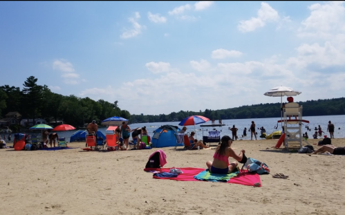 A busy beach scene with people sunbathing, colorful umbrellas, and tents by the water on a sunny day.