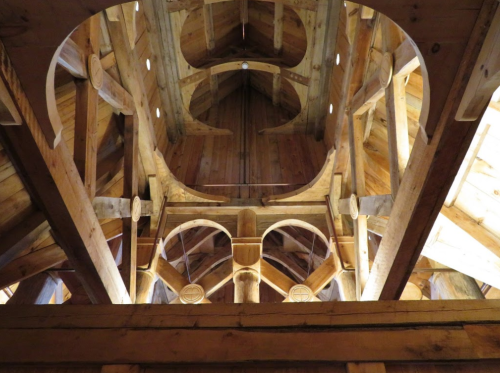 Interior view of a wooden structure showcasing intricate beams and arches against a warm, natural wood backdrop.
