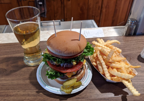A tall burger with lettuce, tomato, and pickles, served with crinkle-cut fries and a glass of beer.