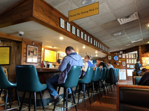 A cozy restaurant interior with patrons seated at a bar, wooden decor, and framed photos on the walls.
