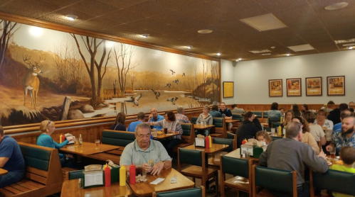 A busy restaurant with wooden tables, patrons dining, and a mural of a serene landscape on the wall.