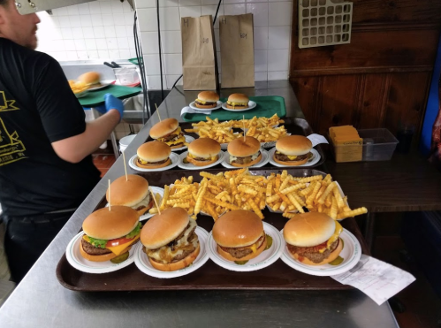 A tray of burgers and fries in a kitchen, with a cook preparing more burgers in the background.