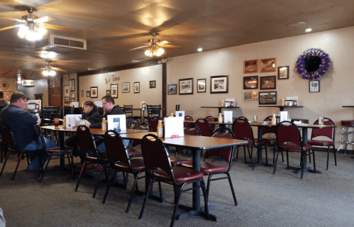 A cozy restaurant interior with empty tables, chairs, and wall decor, featuring a few patrons at a table.