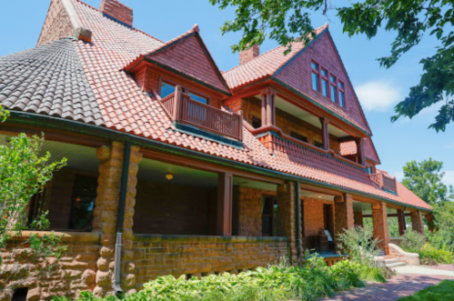 A large, red brick house with a distinctive roof and balconies, surrounded by greenery under a clear blue sky.