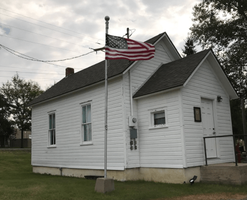 A small white house with a flagpole displaying the American flag in front, surrounded by grass and trees.