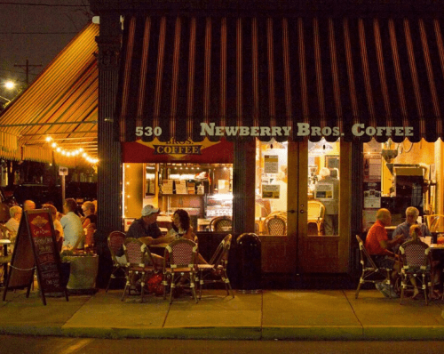 A cozy coffee shop at night, with outdoor seating and warm lights illuminating patrons enjoying their drinks.