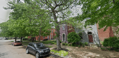 A tree-lined street with parked cars and colorful houses, featuring greenery and a peaceful neighborhood atmosphere.