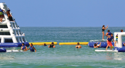 Children and adults play on inflatable water structures in a clear blue ocean under a sunny sky.