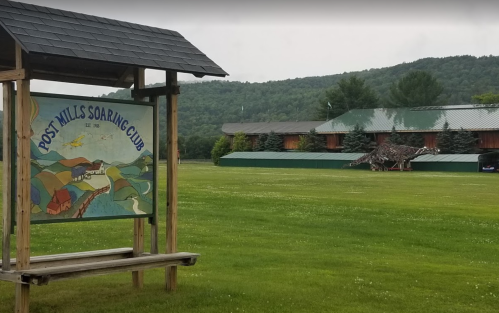 Sign for Post Mills Soaring Club with a scenic background of hills and a building, set in a grassy area.