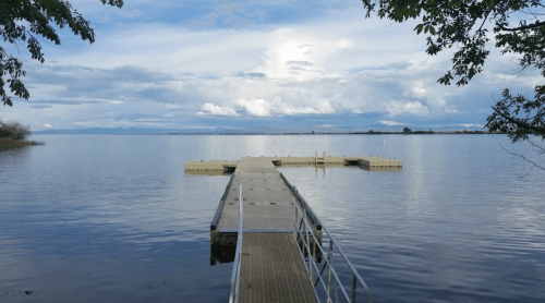 A wooden dock extends into a calm lake, surrounded by trees and under a cloudy sky.